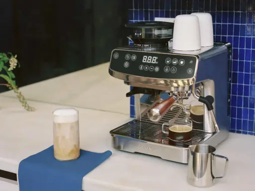 Un angolo caffè casa con macchina espresso, macinacaffè, tazzine e un bicchiere di cappuccino.