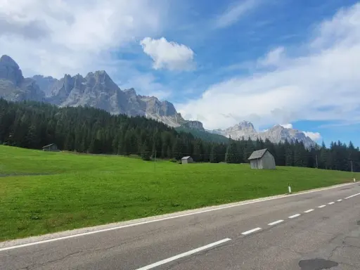 Strada panoramica verso il passo Monte Croce, con prati verdi, boschi di conifere e imponenti vette rocciose sotto un cielo azzurro con nuvole bianche.