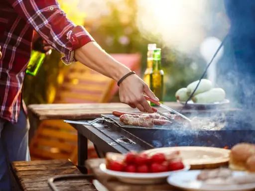 Hombre asa salchichas envueltas en tocino, ideal para saber qué llevar a una barbacoa para sorprender. Hay cervezas y tomates en la mesa.