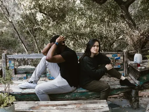 Pareja sentada de espaldas en un banco, con expresiones de angustia. El hombre se agarra la cabeza, ella mira al vacío. Un **vínculo traumático** parece separarlos en este entorno natural.