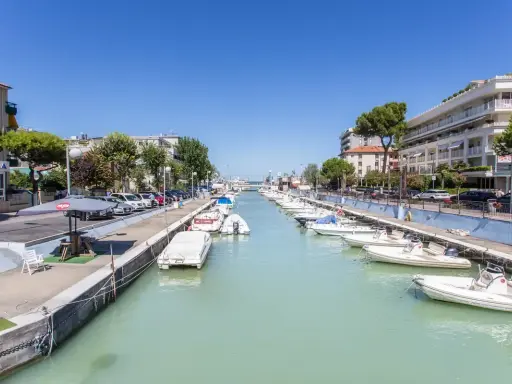 La darsena di Riccione, con barche ormeggiate lungo il canale, palazzi e alberi verdi sotto un cielo azzurro.