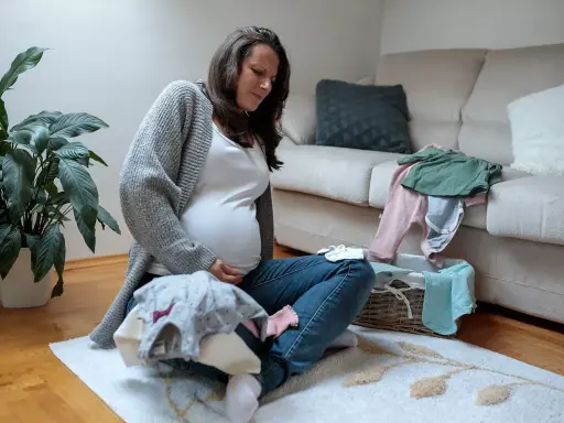 Schwangere Frau sitzt auf dem Boden, hält ihren Bauch und scheint Braxton Hicks Kontraktionen zu spüren, während sie Babykleidung sortiert.