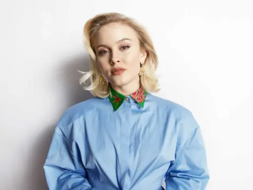 Zara Larsson, känd för sin musik, bär en blå skjorta med en färgglad krage. Hennes skivbolag har säkert hjälpt henne nå framgång.