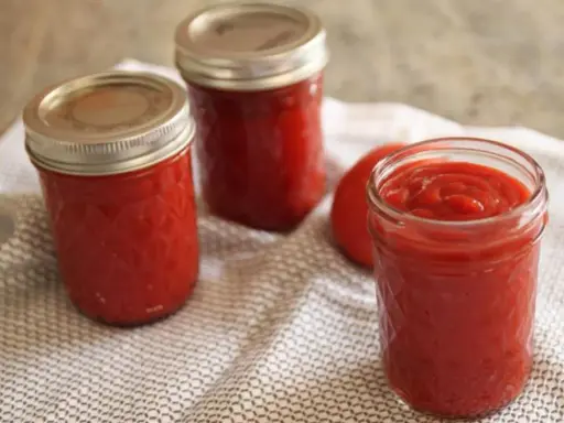 Trois bocaux de ketchup maison, un rouge vif, reposent sur une serviette à motifs. Un tomate entière est visible.