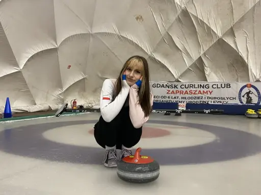 Młoda sportsmenka na lodzie, gotowa do gry w curling. W tle baner "Gdańsk Curling Club Zapraszamy".