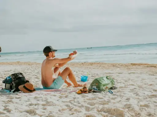 Mężczyzna robi zdjęcie telefonem na plaży, marząc o najkrótszych lotach z Polski na wakacje. Obok leżą torba, kapelusz i sandały.