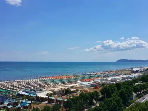 Ampia spiaggia di Riccione con file ordinate di ombrelloni e lettini, bagnanti che si godono il sole e l'acqua cristallina del mare Adriatico sotto un cielo azzurro.