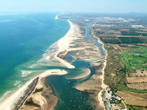 Luftaufnahme der Ria Formosa mit ihren Sandbänken, Lagunen und dem Meer. Im Hintergrund sind Felder und ein Golfplatz zu sehen.
