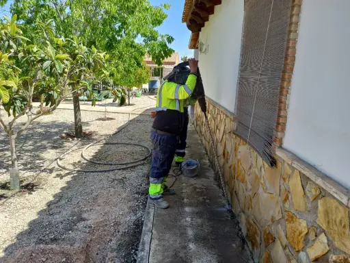 Trabajador con equipo de protección realiza trabajos de inyección de cemento en la base de una pared exterior, rodeado de vegetación.