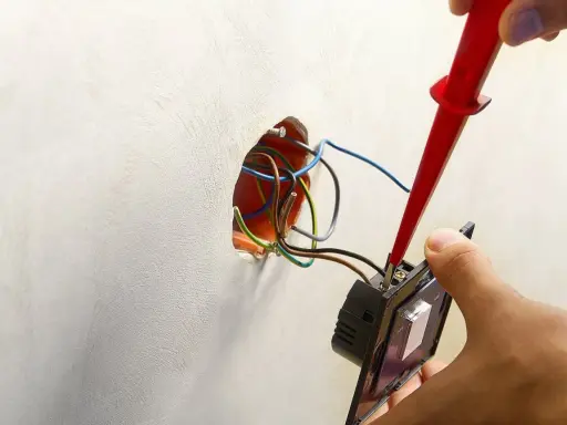 Ręce montują włącznik światła, podłączając przewody. Widać instalację elektryczną i narzędzie.