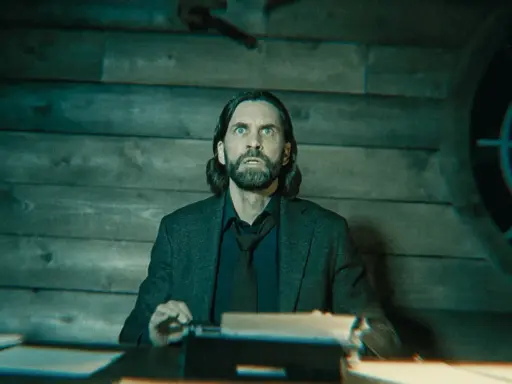 Alan Wake 2: un hombre con barba y pelo largo, con expresión de sorpresa, sentado frente a una máquina de escribir en una habitación de madera.