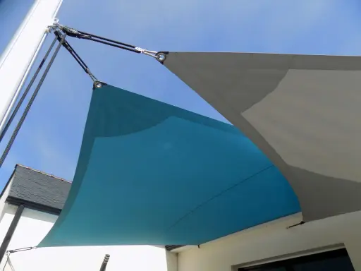 Comment installer un voile d'ombrage sur terrasse : deux voiles, bleu et gris, fixés par des cordages et des poulies à un mât et à la maison.