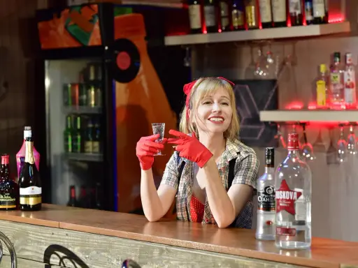 Uśmiechnięta barmanka w czerwonych rękawiczkach trzyma kieliszek. Na ladzie stoi duża butelka wódki, idealna na pół litra na dwóch.