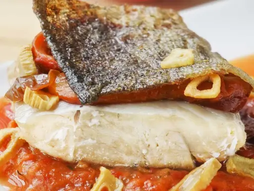 Bacalao a la riojana con piel crujiente, pimientos y ajo frito, servido sobre una cama de salsa de tomate.