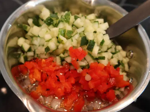 Finhackad gurka och röd paprika i en kastrull, redo för ett bostongurka recept. En sked vilar i blandningen.