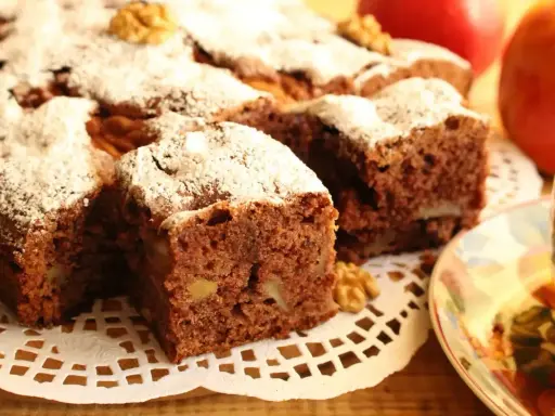 Kawałki aromatycznego ciasta cynamonowego z orzechami, posypane cukrem pudrem, obok jabłek i filiżanki herbaty.