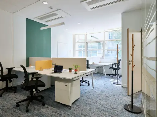 Un bureau chaleureux avec des postes de travail modernes, des cloisons acoustiques et un tapis bleu doux. L'espace est lumineux grâce aux grandes fenêtres.