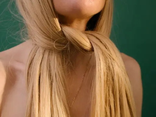 Blondes, gesundes Haar, das sich um den Hals schlingt. Ein zarter goldener Anhänger ist unter den Haaren zu sehen.