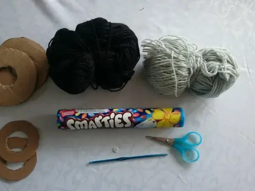 Materialien für ein Geldgeschenk: Pappringe, schwarzes und hellgraues Garn, Smarties-Rolle, Knopf, Häkelnadel und Schere. So lässt sich eine Hantel basteln.