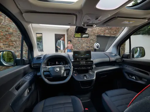 Interior de la nueva Citroën Berlingo, con pantalla táctil, volante moderno y asientos cómodos. Afuera, una pareja se acerca a un punto de carga.