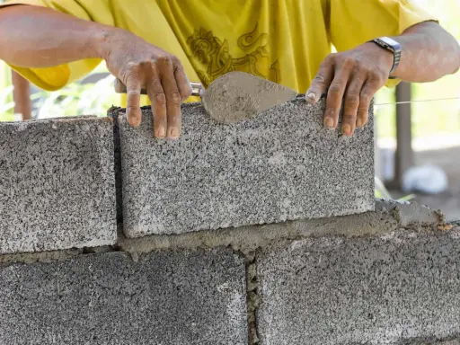 Budowa fundamentu z bloczków betonowych. Ręce murarza układają kolejny bloczek, używając kielni z zaprawą.