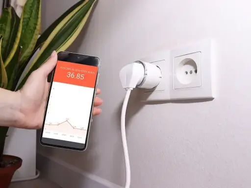 Smart wtyczka w gniazdku i smartfon z wykresem pokazującym monitoring zużycia energii.