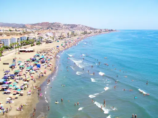 Ein belebter Strand in Rincón de la Victoria, mit vielen Sonnenschirmen, Menschen am Wasser und im Meer.