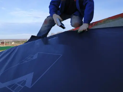 Un ouvrier en bleu, ganté, utilise un outil pour la pose d'un pare pluie sur un toit. Le ciel est bleu avec quelques nuages.