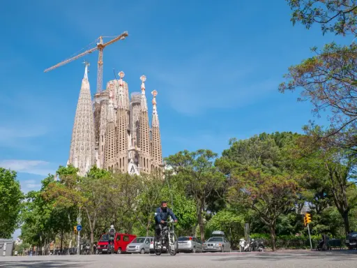 Sagrada Familia w Barcelonie, wciąż w budowie, góruje nad zielonym parkiem. Na ulicy widać rowerzystę i samochody.