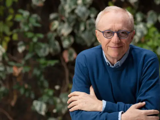 David Grossman, con occhiali blu e maglione scuro, sorride con le braccia incrociate su sfondo verde.