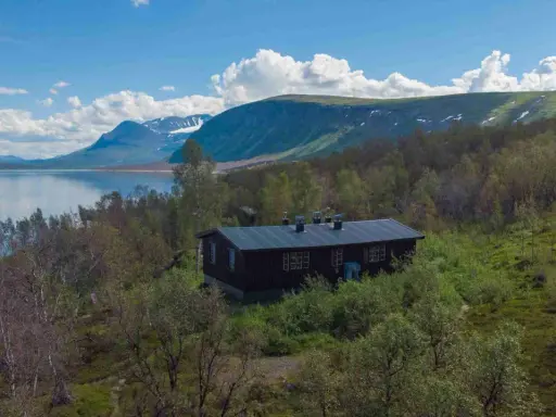 Mörkbrun byggnad vid en sjö, omgiven av björkskog och med fjäll i bakgrunden. En idyllisk plats för ritsem camping.