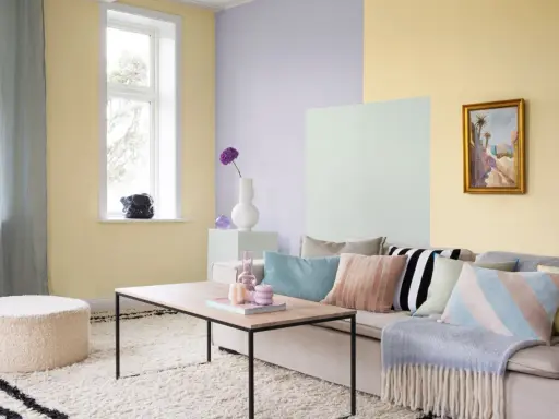 Ett vardagsrum med pastell inredning. Ljusgula, ljuslila och ljusgröna väggar skapar en mjuk atmosfär. En beigefärgad soffa är fylld med kuddar i olika pastellfärger och mönster.