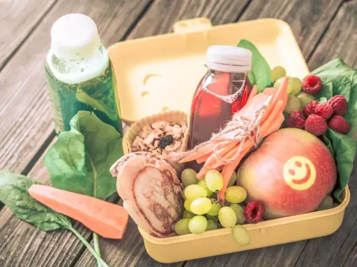 Zdrowa dieta 3 latka: żółty lunchbox z jabłkiem, malinami, winogronami, marchewką, placuszkiem i sokami.