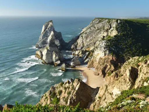 Steile Felsklippen umgeben einen einsamen sintra strand. Das türkisfarbene Meer bricht sich an den Felsen.