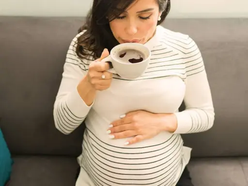 Schwangere Frau trinkt eine Tasse entkoffeinierten Kaffee, während sie ihren Bauch streichelt.