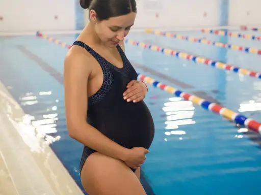 Kobieta w ciąży w czarnym stroju kąpielowym stoi przy basenie, delikatnie dotykając brzucha.