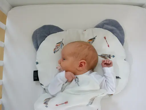 Nowe niemowlę śpi na poduszce w kształcie misia, otulone kocykiem z piórkami. Czy niemowlę może spać na poduszce? Bezpieczeństwo przede wszystkim!