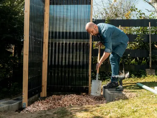 Uomo costruisce una doccia da giardino fai da te, livellando ghiaia con una pala.