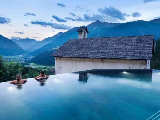 Due donne in una piscina a sfioro ammirano il panorama di Bormio e dintorni, con montagne maestose e un piccolo edificio storico.