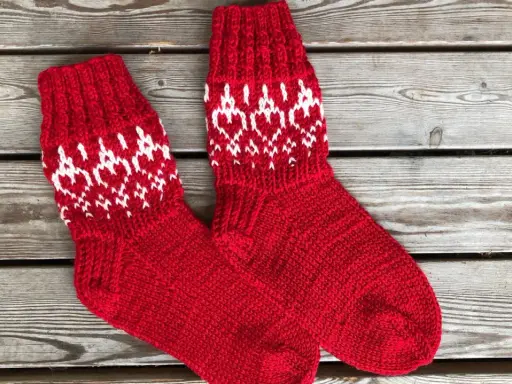 Två par röda, stickade strumpor med vitt mönster av hjärtan och snöflingor ligger på en träbänk. Perfekta för kalla dagar!