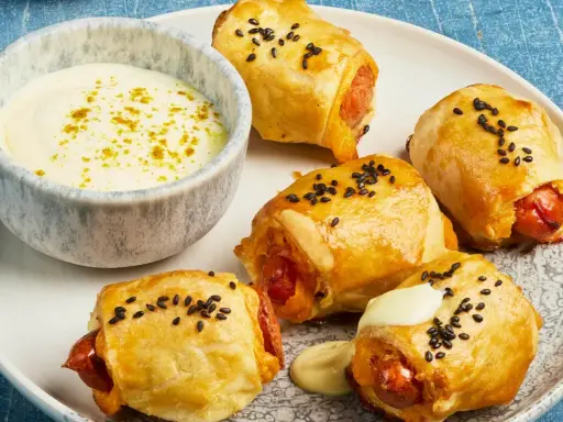 Goldbraune Blätterteigkissen mit Würstchen, bestreut mit schwarzen Sesamkörnern, serviert mit einer cremigen Dip-Sauce.
