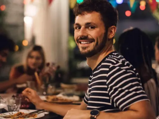 Uśmiechnięty mężczyzna na tle przyjaciół przy stole. Idealny, tani pomysł na wieczór kawalerski z jedzeniem i dobrym towarzystwem.