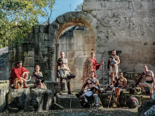 Un groupe de guerriers, prêts à combattre, pose devant des ruines antiques. Ces hommes, tels des gladiateurs romains, arborent armures et armes, prêts pour l'arène.