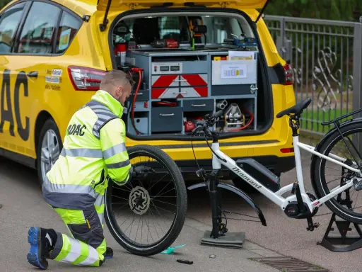 ADAC-Mitarbeiter repariert eine Fahrradpanne. Ein gelbes ADAC-Auto mit Werkzeugkasten steht daneben.