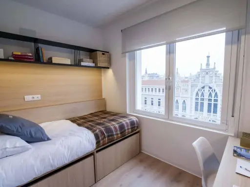 Habitación moderna con cama, estanterías y vistas a un edificio histórico en Xior Madrid Retiro.