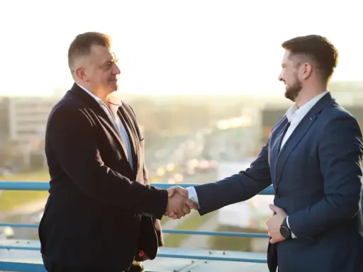 Dwóch mężczyzn w garniturach ściska sobie dłonie na tle miasta.