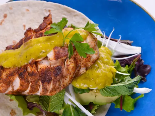 Delicioso wrap de pollo con aguacate, lechugas variadas y salsa verde, servido en un plato azul vibrante.