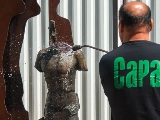 Hombre con camiseta negra con letras verdes "Capa" limpia una estatua de bronce con una manguera a presión.