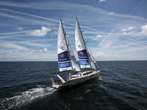 Le voilier Pangaea de Mike Horn navigue sur une mer calme sous un ciel bleu parsemé de nuages et de traînées d'avions.
