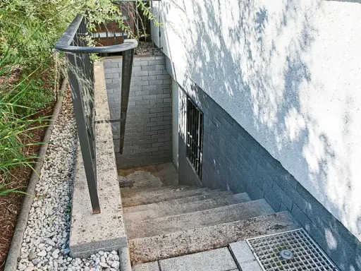 Betontreppe sanieren: Alte Steintreppe mit Geländer führt zu einer Kellertür. Grüne Pflanzen und Kies säumen den Weg.